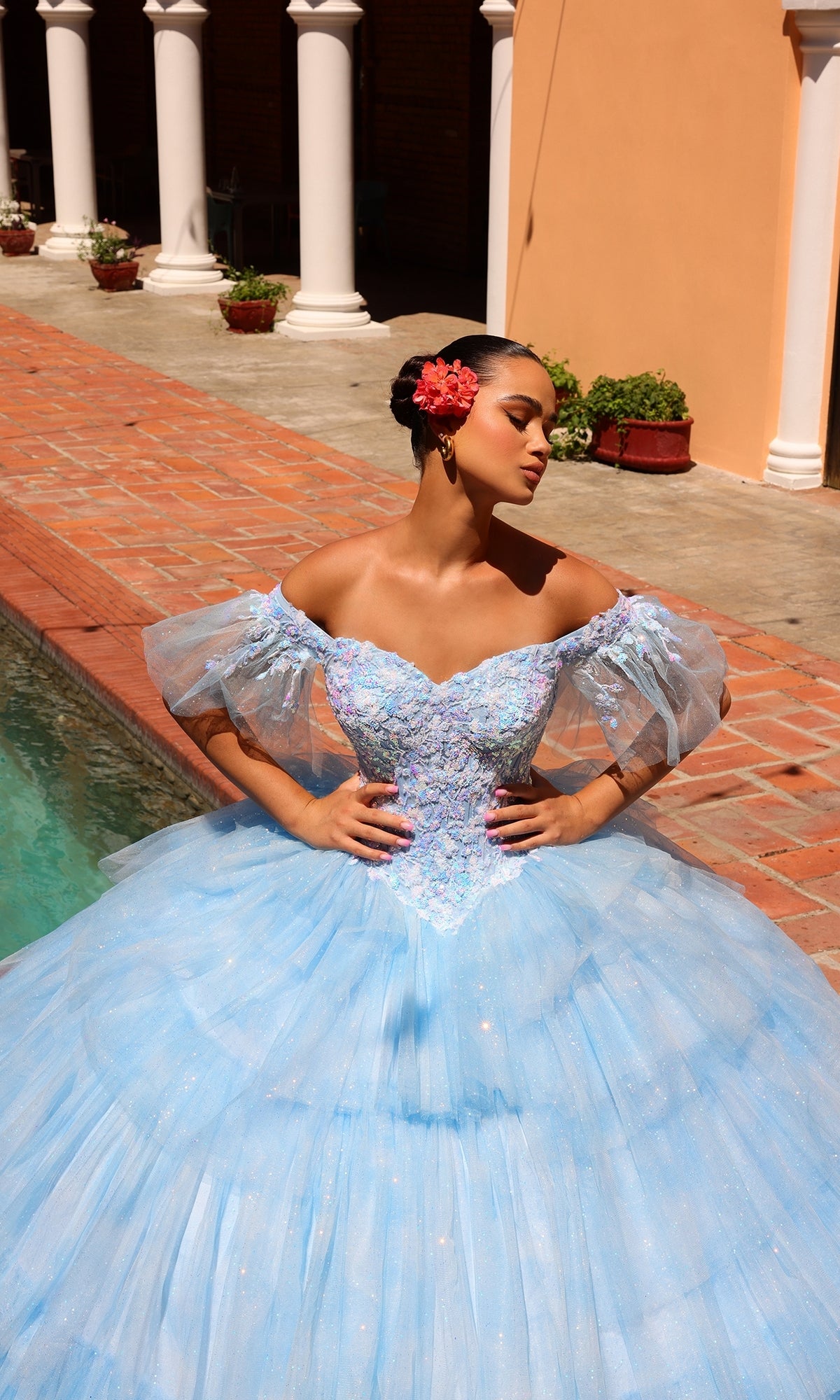 A woman in the Quinceanera Dress 54312 By Amarra?a blue off-the-shoulder ball gown with puffed sleeves and a sweetheart neckline?stands by a pool, hands on hips, with a red flower in her hair.